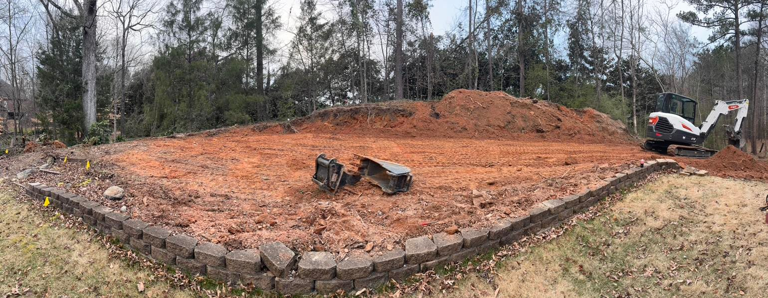 A construction site with two mini-excavators moving dirt. A retaining wall borders the site, surrounded by trees.