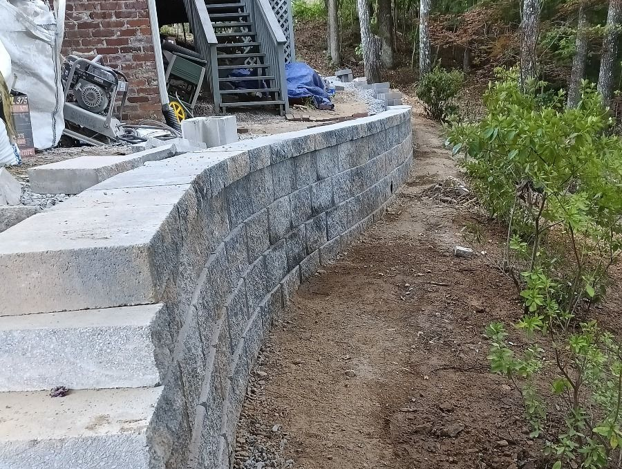 Stone retaining wall curving along a dirt path, with stairs and a building in the background.