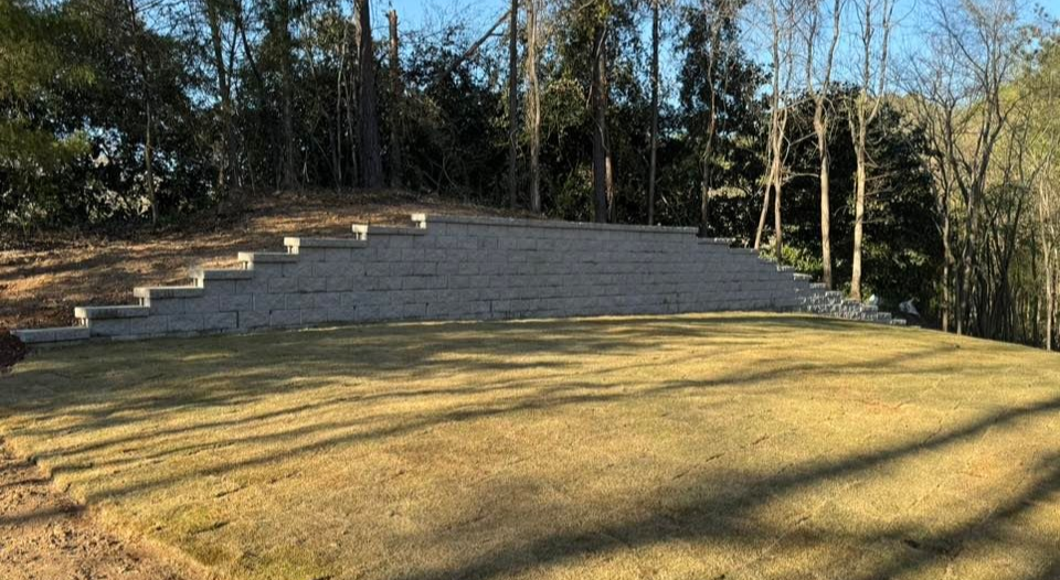 Grassy backyard with tiered retaining wall and steps leading to trees under a blue sky.