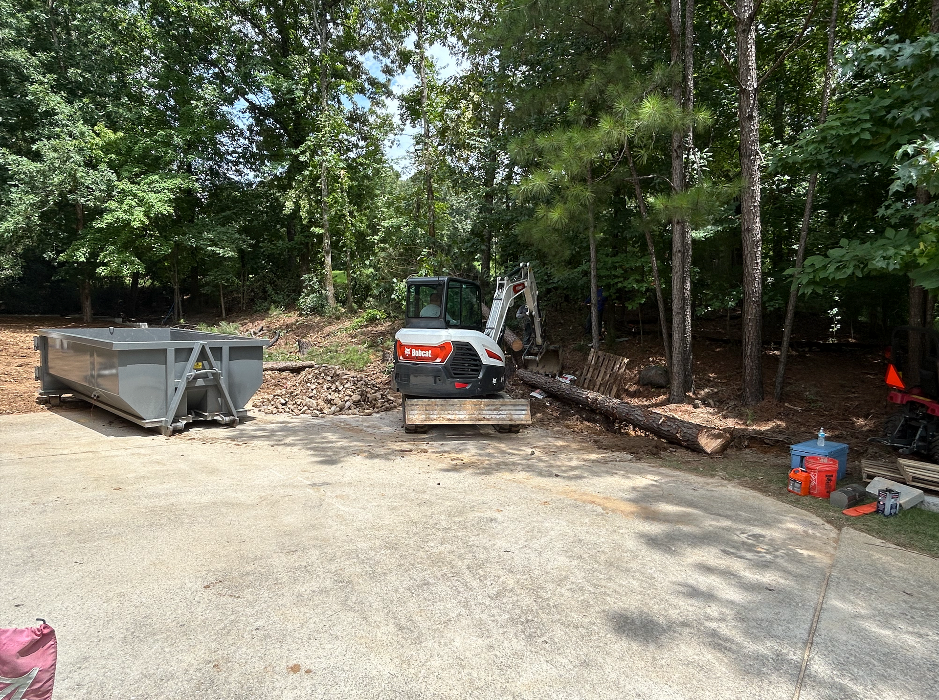 White Bobcat excavator and skid-steer loader digging in a wooded area, brown soil.