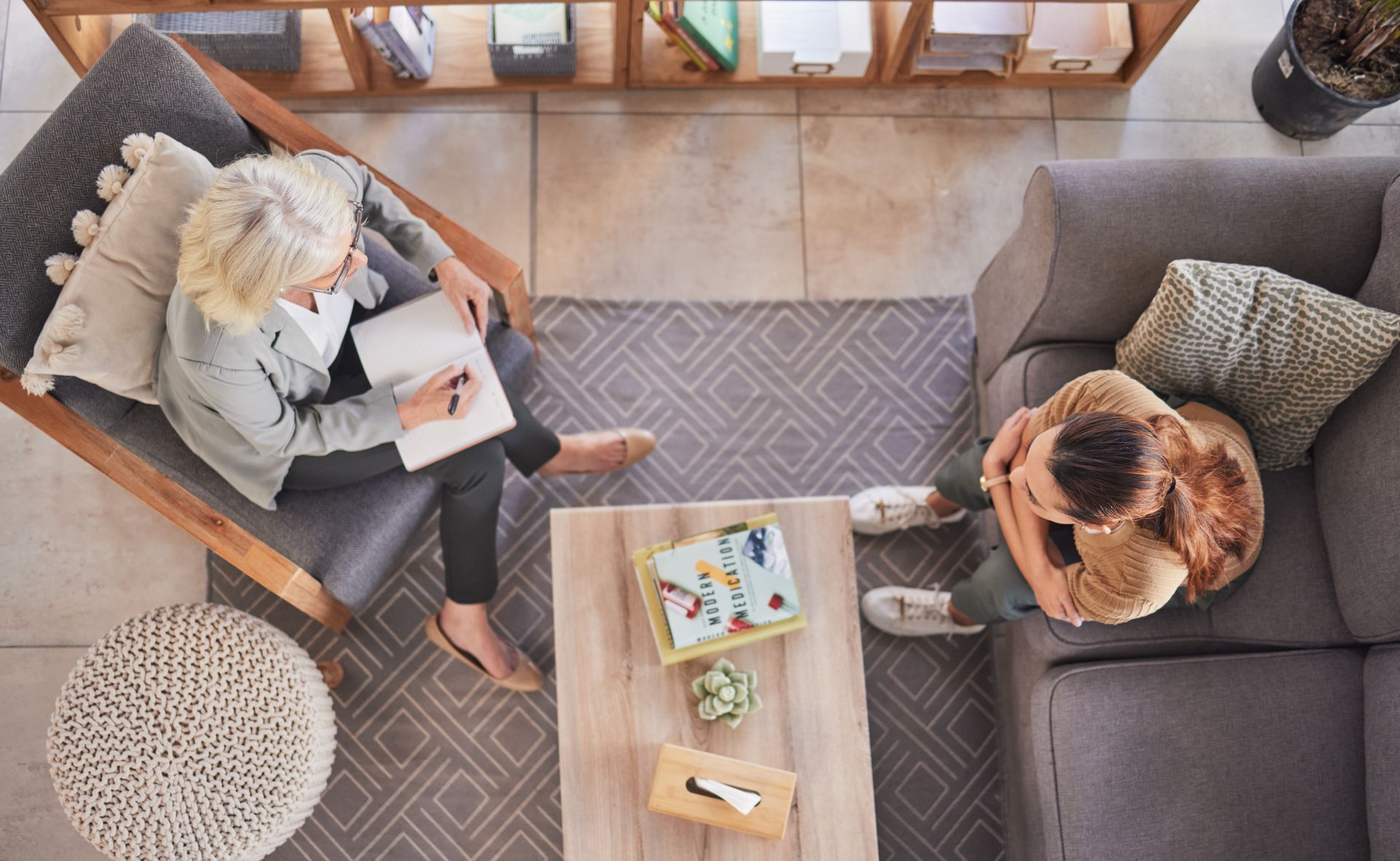 Woman in armchair taking notes; other sits on sofa, face in hands, in therapy session.