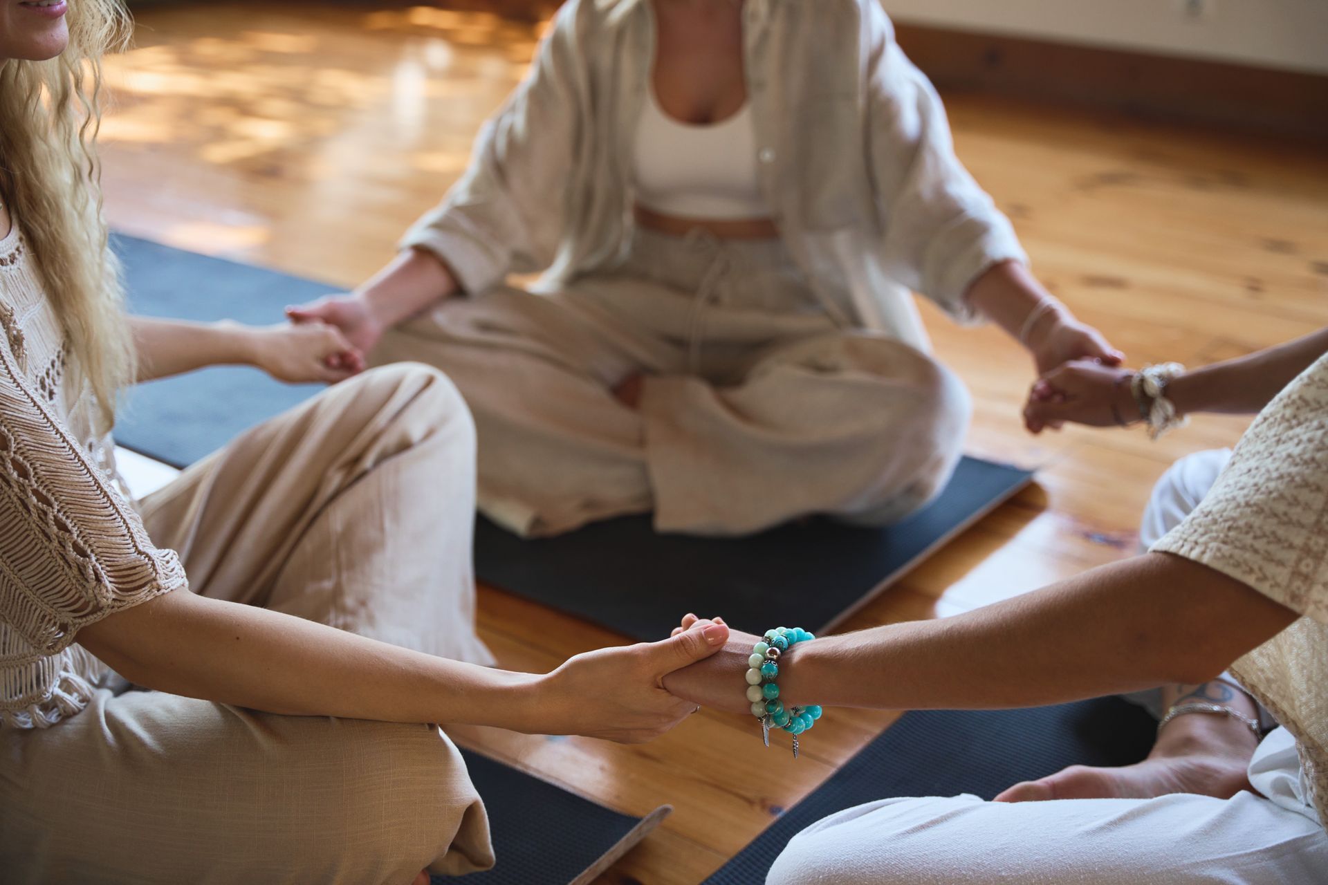 People in a circle, holding hands, meditating on yoga mats. Wooden floor, soft light.