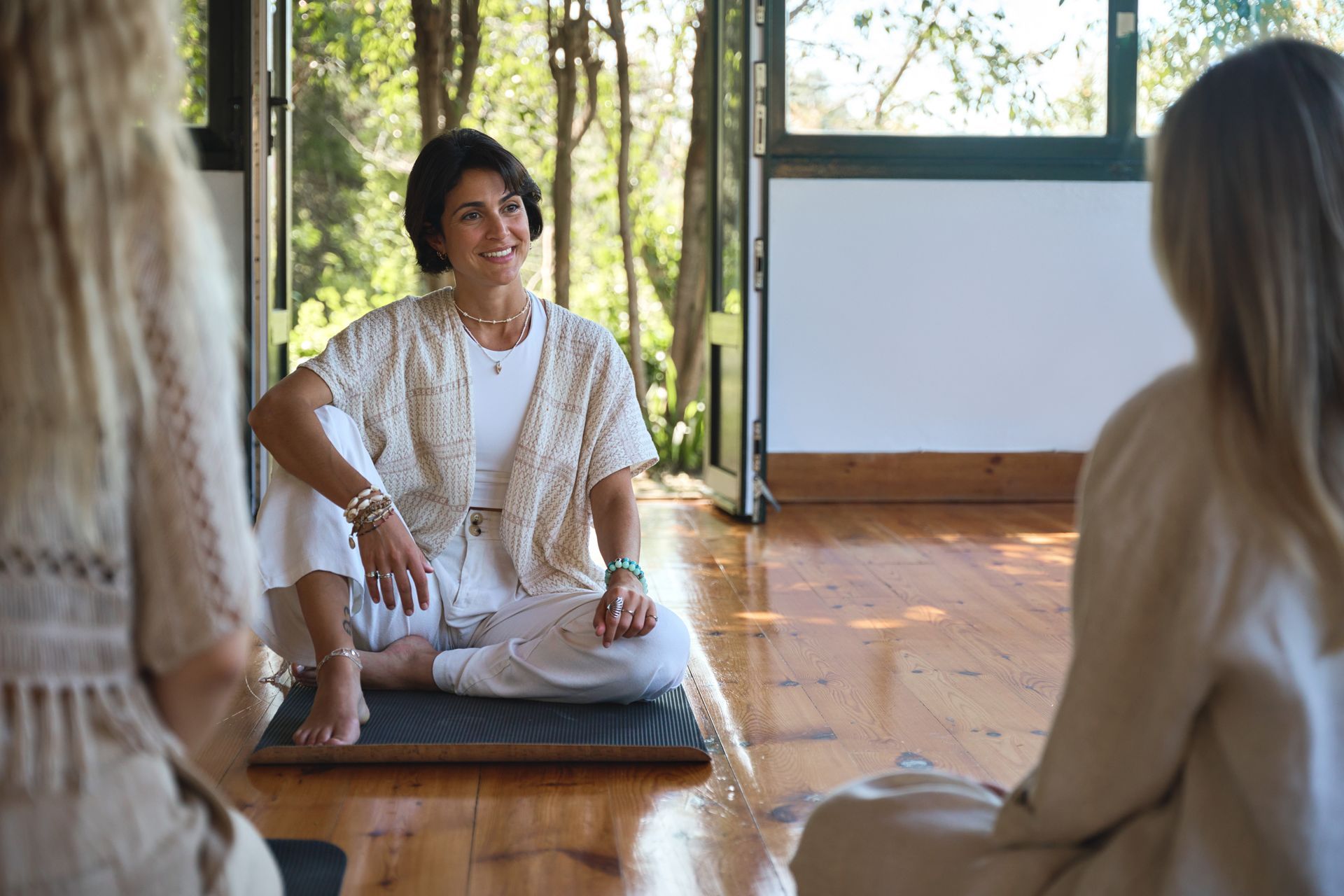 Woman in white outfit smiles while sitting on a mat with two others. Forest setting.