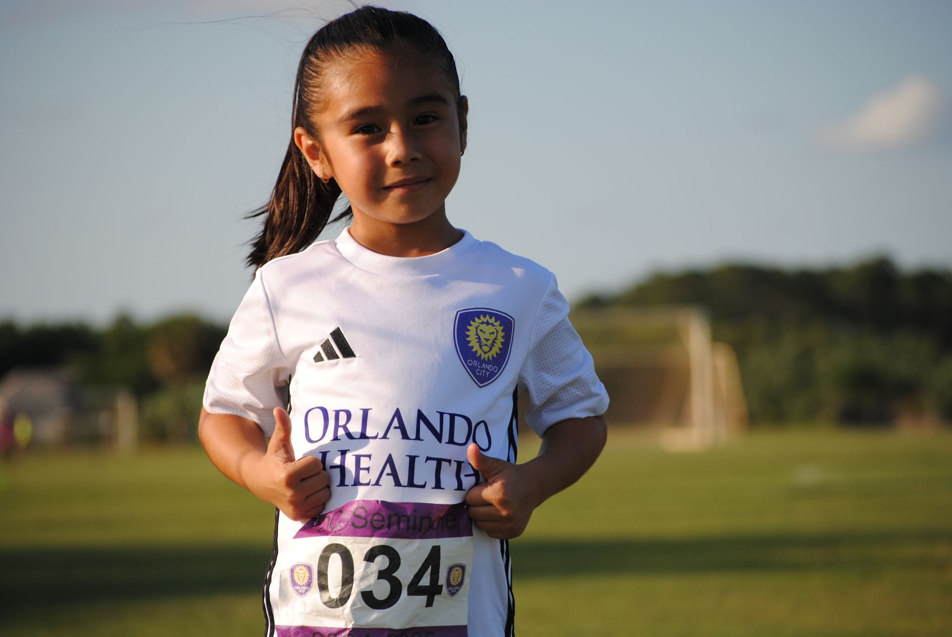 A picture of a little girl holding her thumbs up and dressed in a white soccer uniform