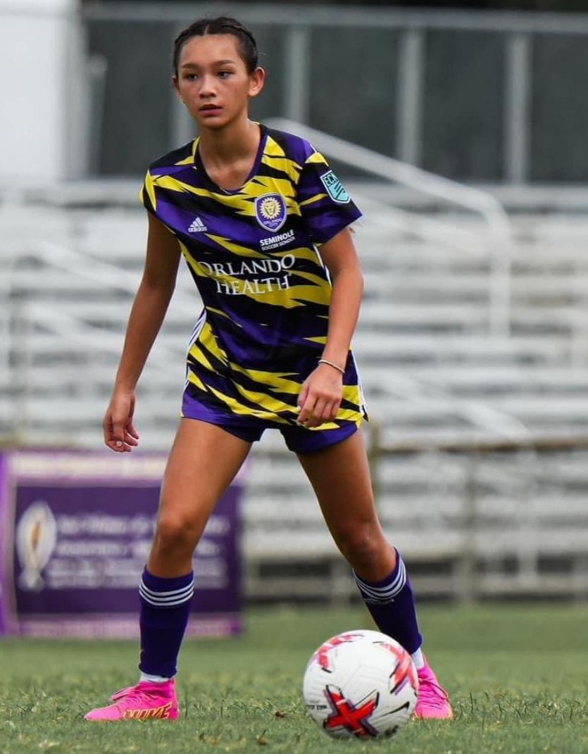 A girl standing with a soccer ball at her feet with the stadium bleachers behind her