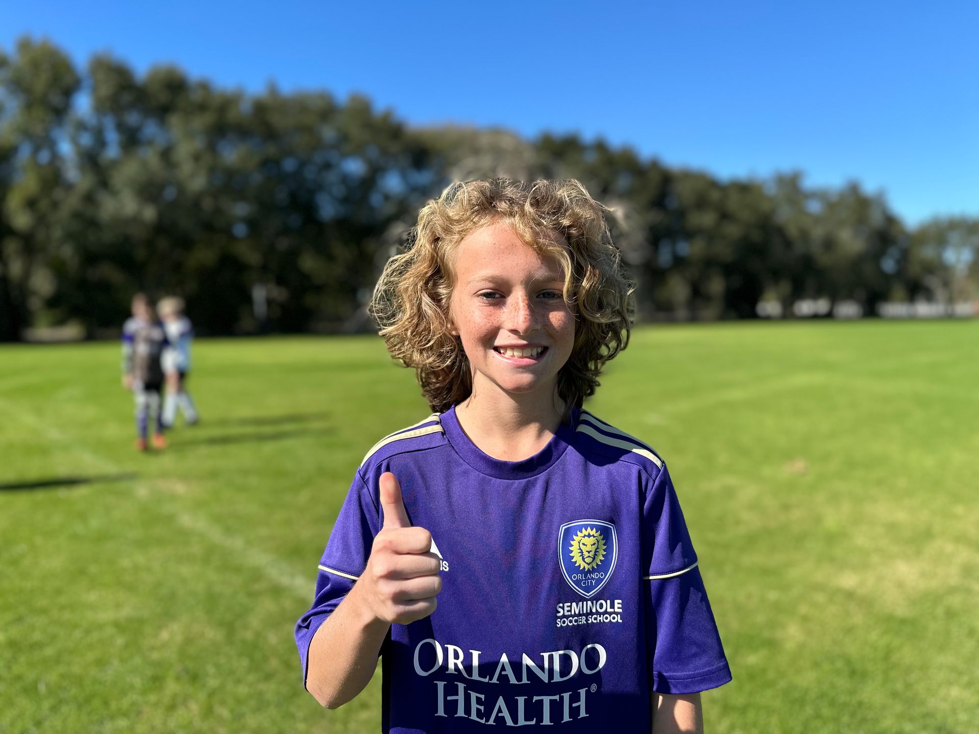 a bonde boy holding his thumb up on the soccer field and smiling