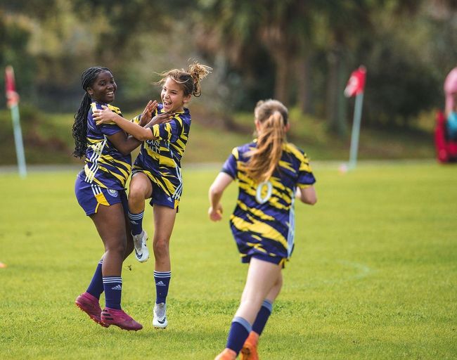 Two girls celebrate as their team just scored a goal. They are hugging one another while another girls runs by.