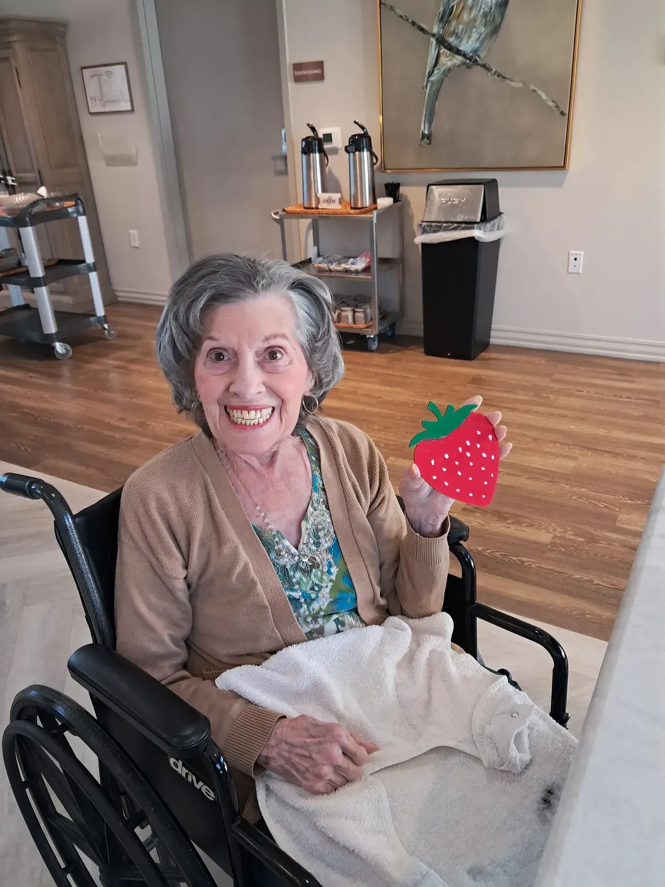 Elderly woman in a wheelchair smiles, holding a strawberry-shaped craft in a bright room.
