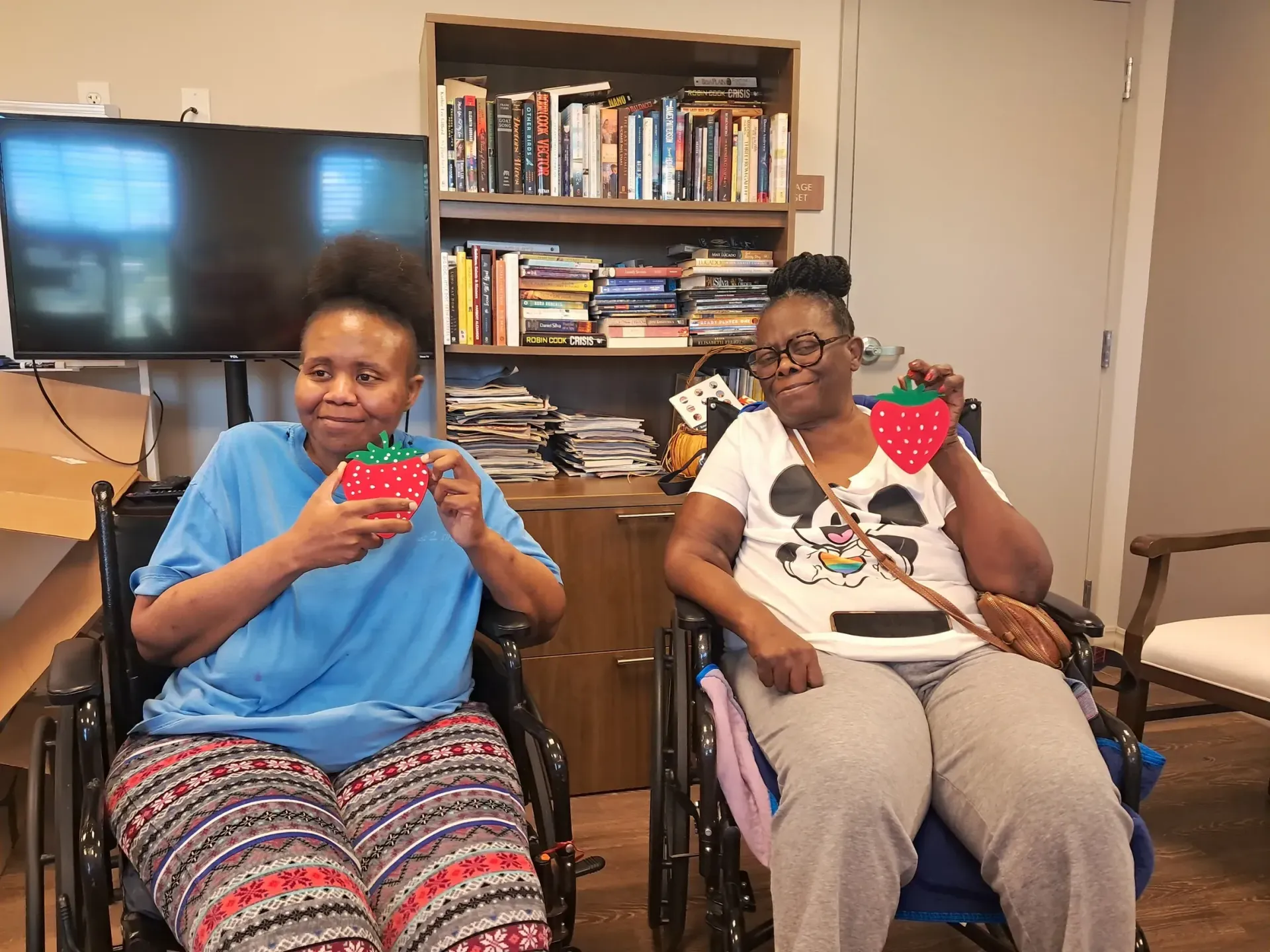 Two women in wheelchairs holding strawberry-shaped crafts, smiling. Indoors, bookcase in the background.