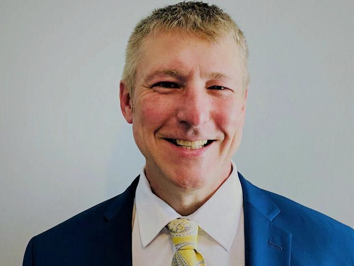 Man in blue suit and yellow tie smiles, looking directly at the camera.
