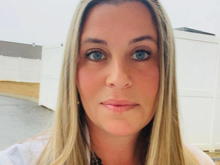 Blonde woman with blue eyes looking at the camera; outdoor setting with white fence in the background.