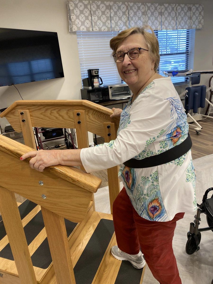 Woman using a stair trainer in a rehab facility, smiling, wearing a safety belt and red pants.