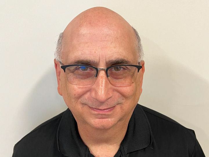 Bald man with glasses, black shirt, smiling in front of a white wall.