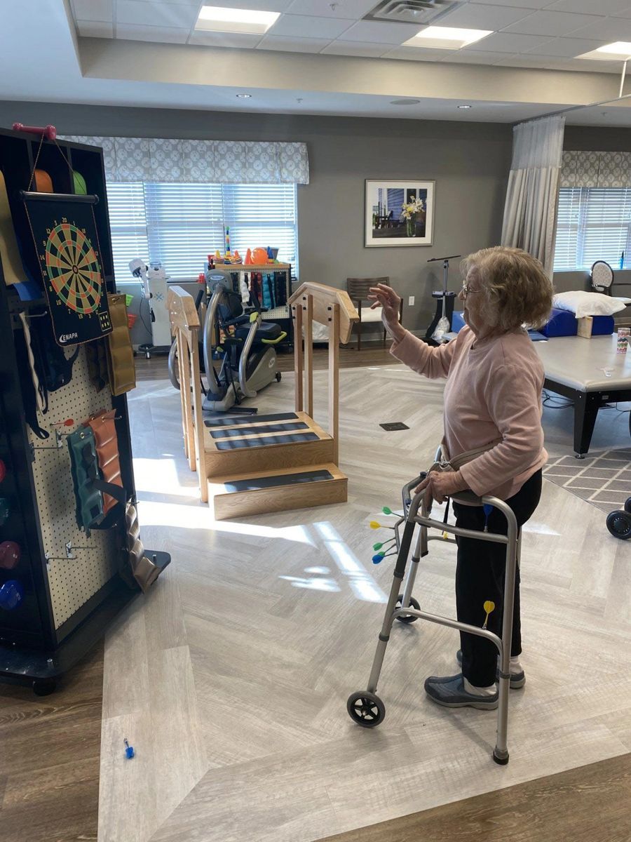 Elderly woman with walker playing darts in a therapy room.