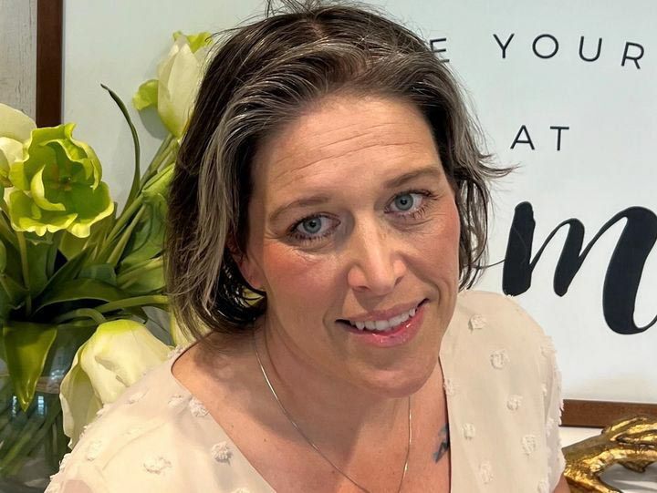 Woman with short, graying hair, smiling, wearing a light-colored top, near flowers and a sign.
