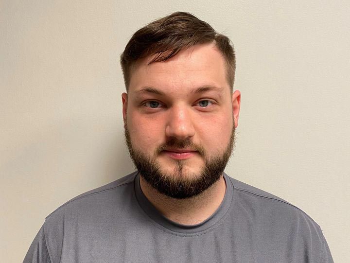 Man with a short beard and haircut, wearing a gray shirt, looking forward against a white wall.