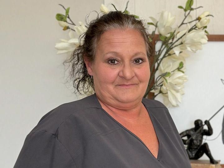 Woman with dark hair in gray shirt smiles; background features flowers, art.