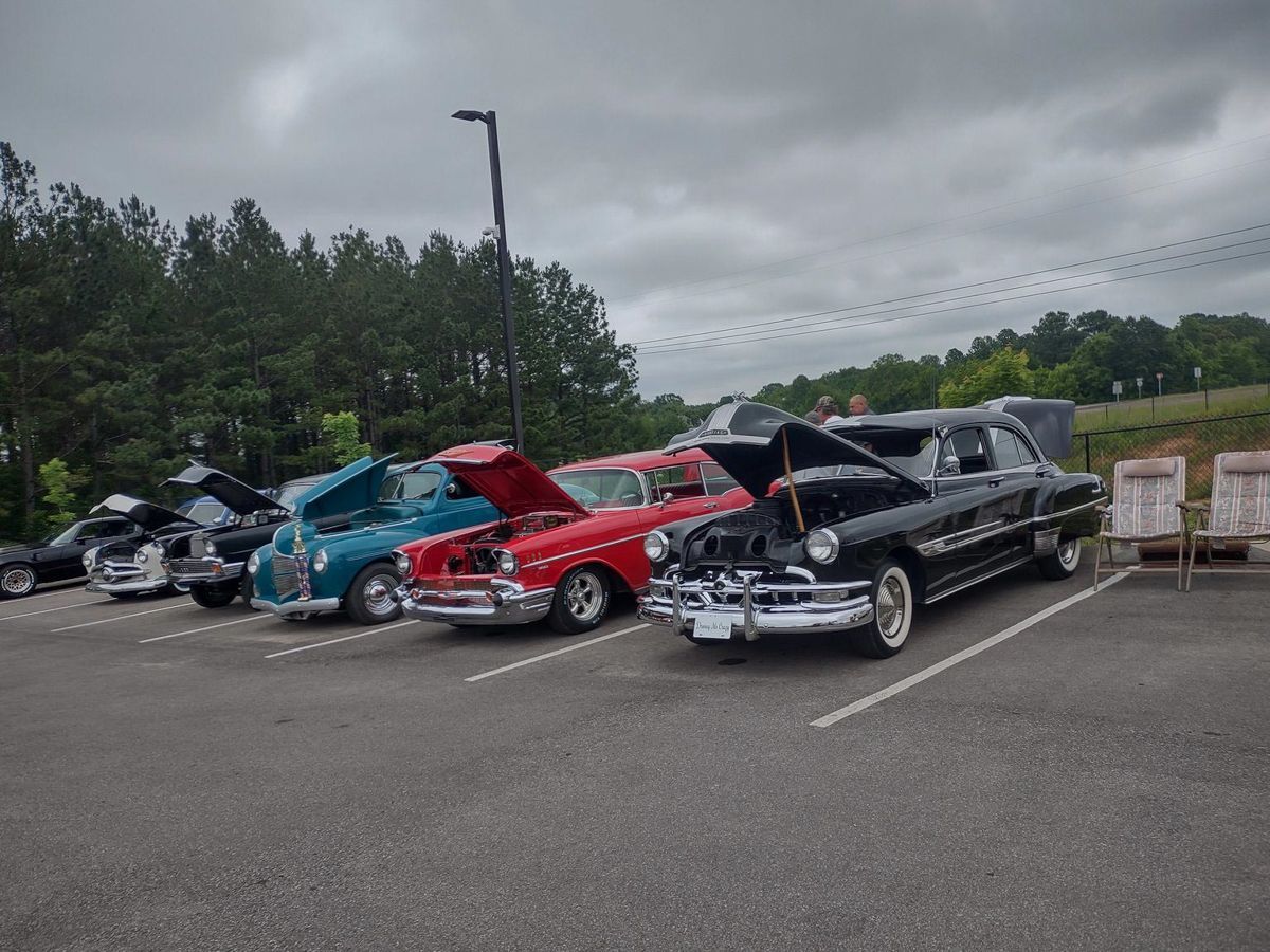 Classic cars parked in a lot, hoods up, with a tree-lined background under a cloudy sky.