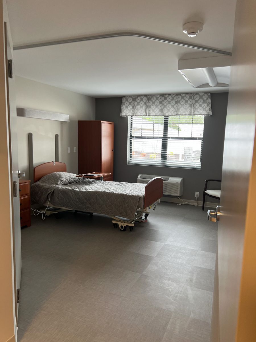 Hospital room: Bed with tray, dresser, window, closet. Gray walls, flooring. View through doorway.