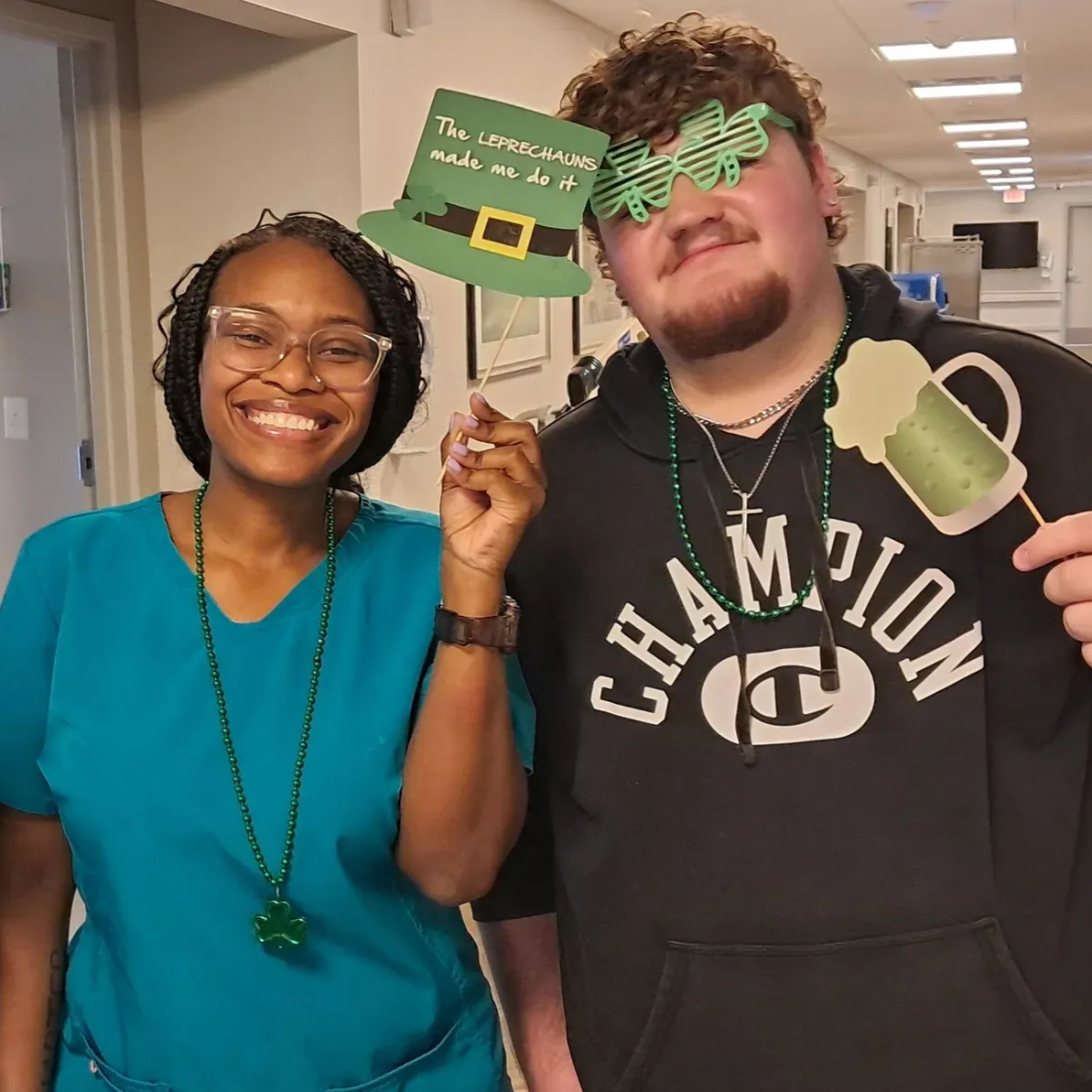 Two people with St. Patrick's Day props: woman in teal scrubs, man in Champion hoodie smiling. Indoors.