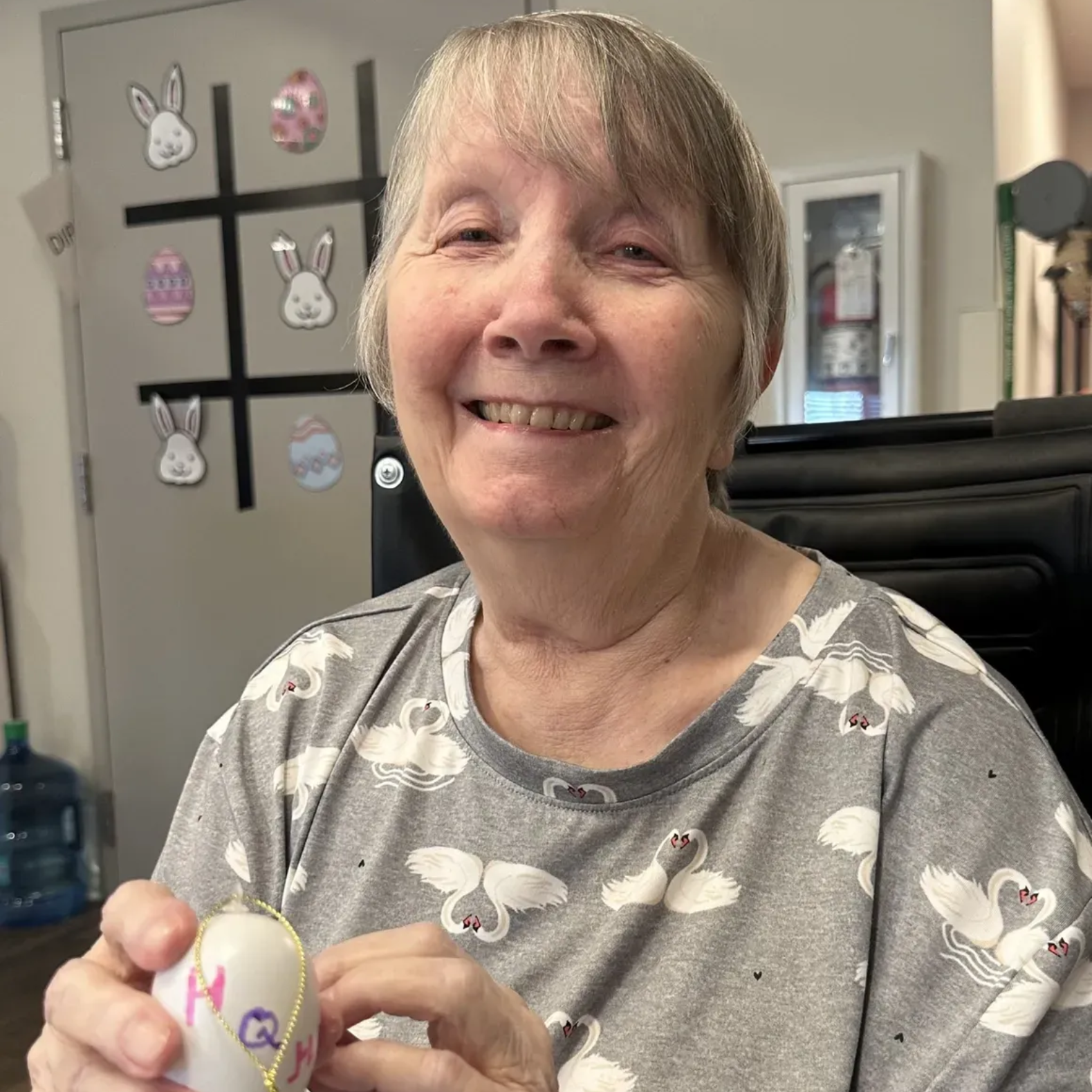 Woman smiling, holding painted Easter egg, gray shirt with swan pattern. Easter-themed door in background.