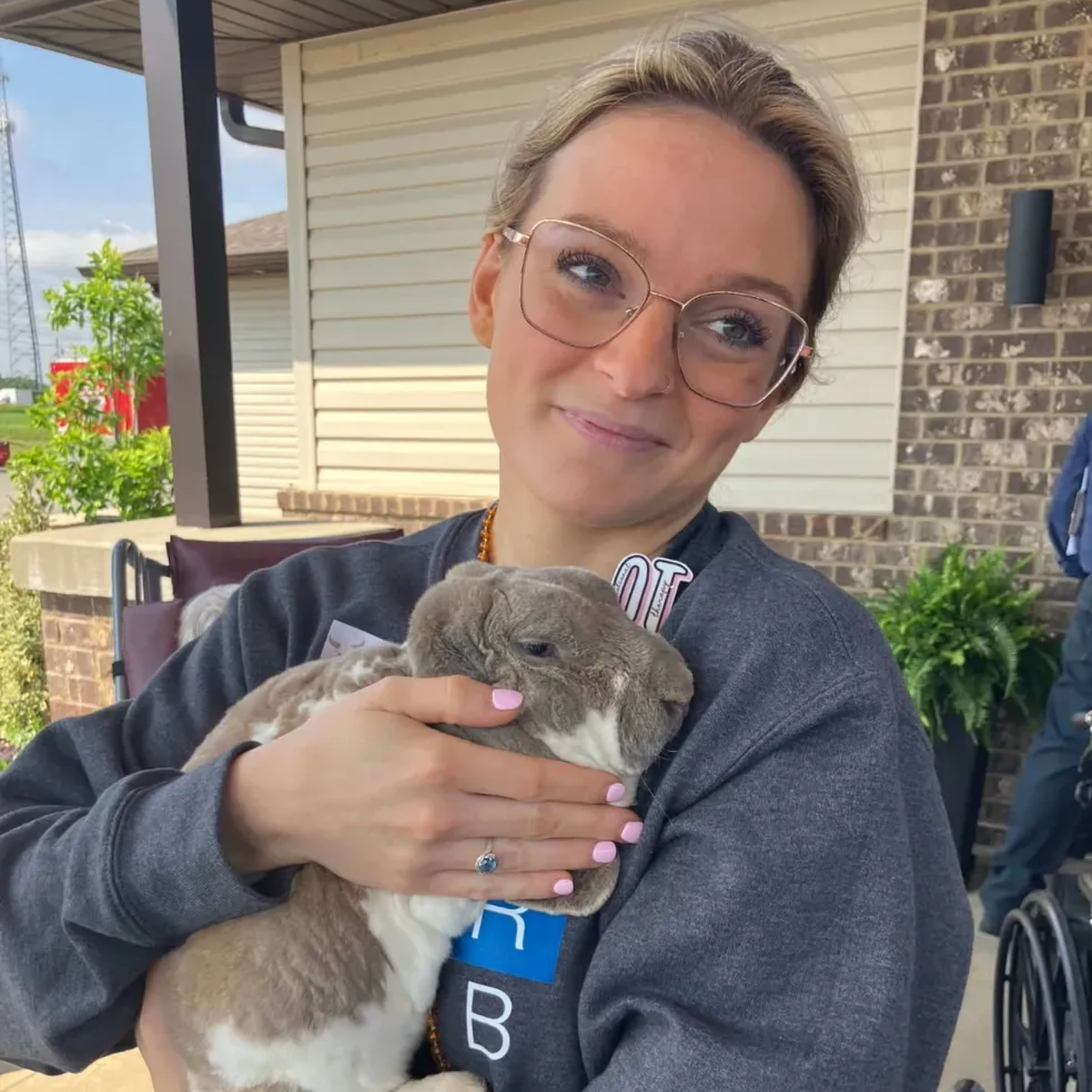 Woman with glasses, smiling, holding a brown and white rabbit, outdoors.