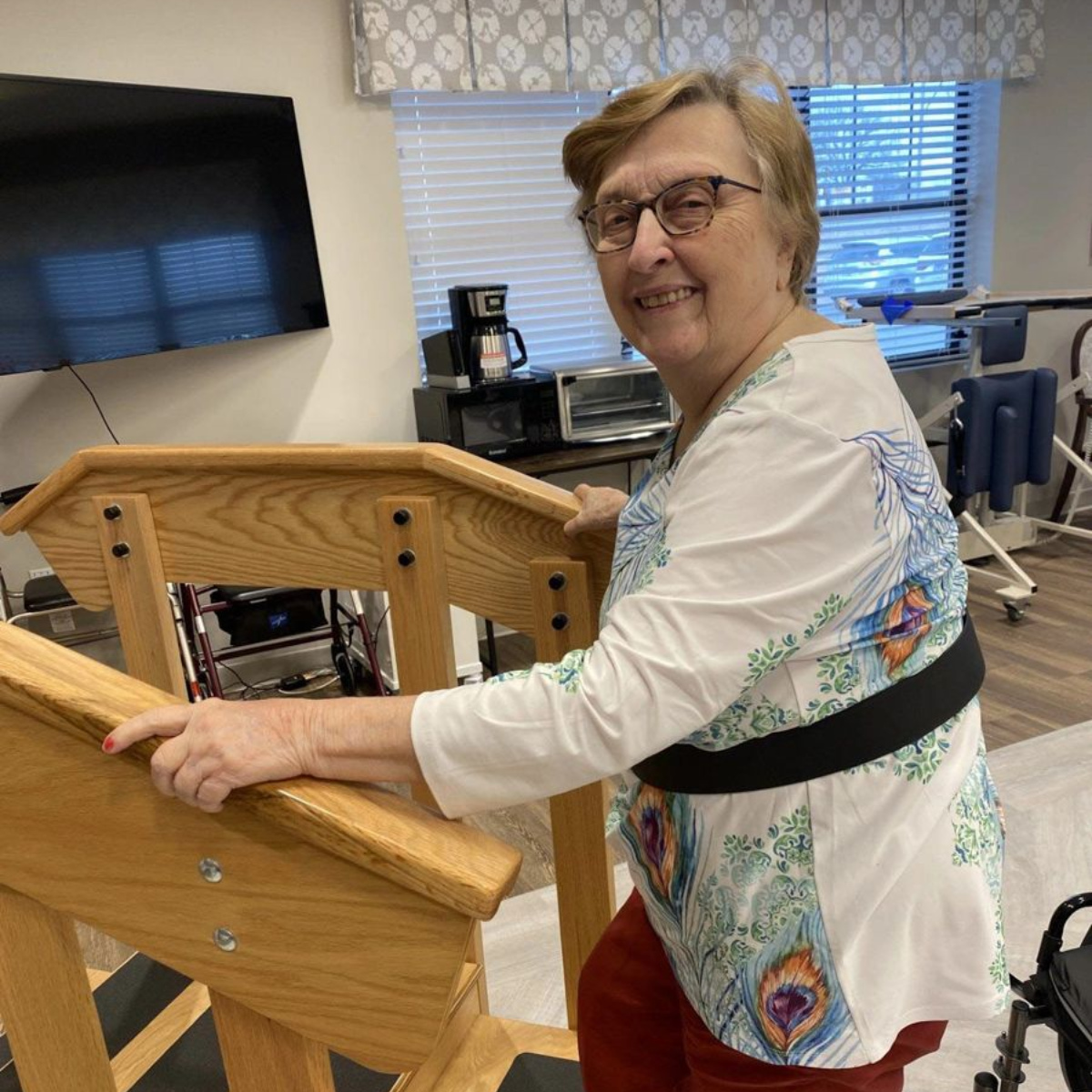 Woman using a wooden staircase for physical therapy, smiling. She's wearing glasses and red pants. Therapy setting.