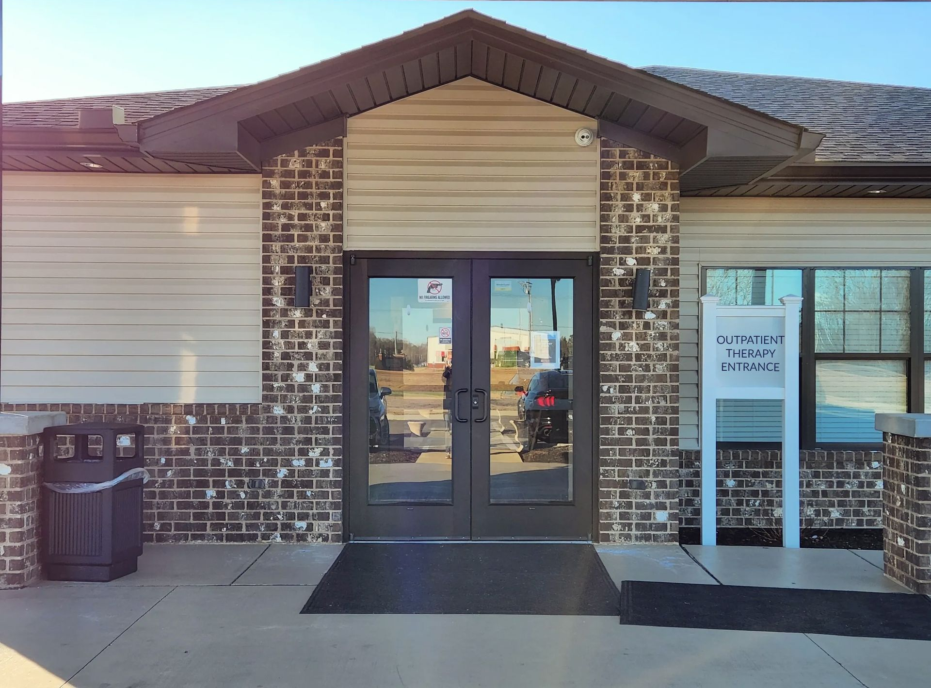 Entrance of a brick building with double doors, trash can, and sign on a sunny day.