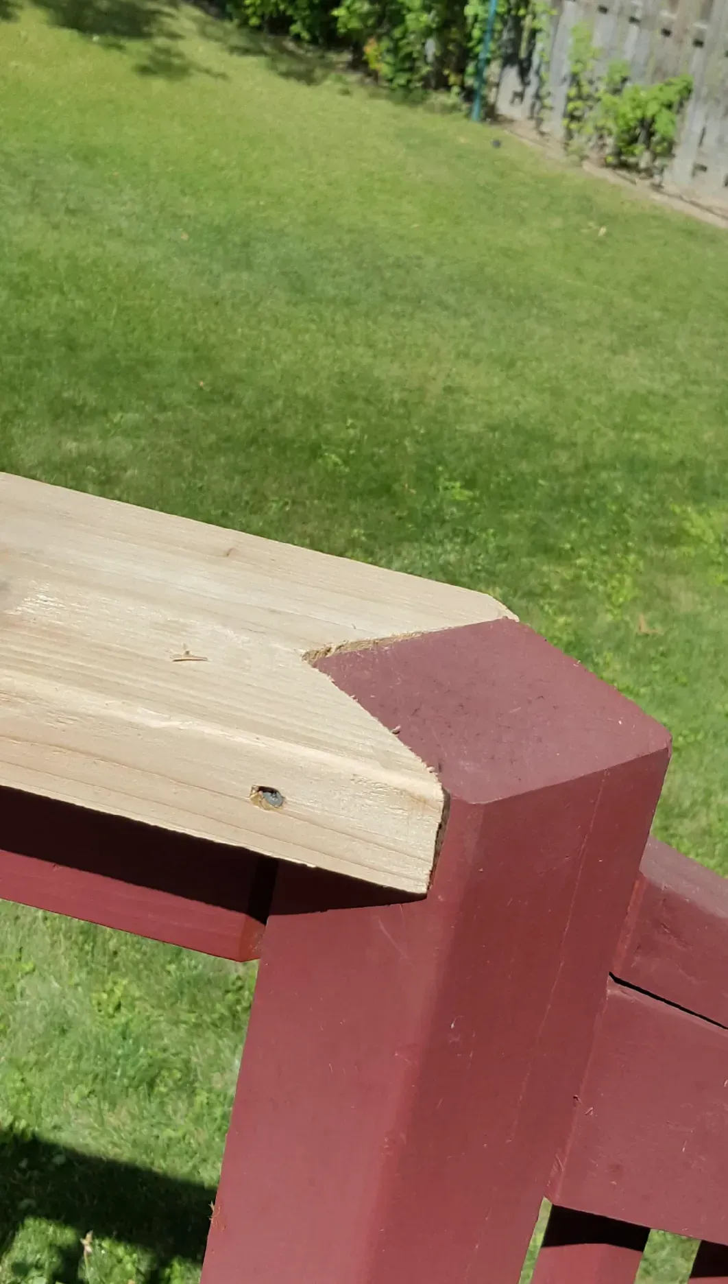 Close-up of a deck railing corner. Light wood meets a maroon-painted post, with a green lawn in the background.