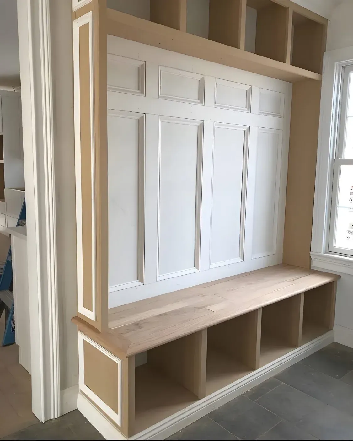 Built-in wooden bench with cubbies, white paneling, and upper shelves, beside a window and wall.
