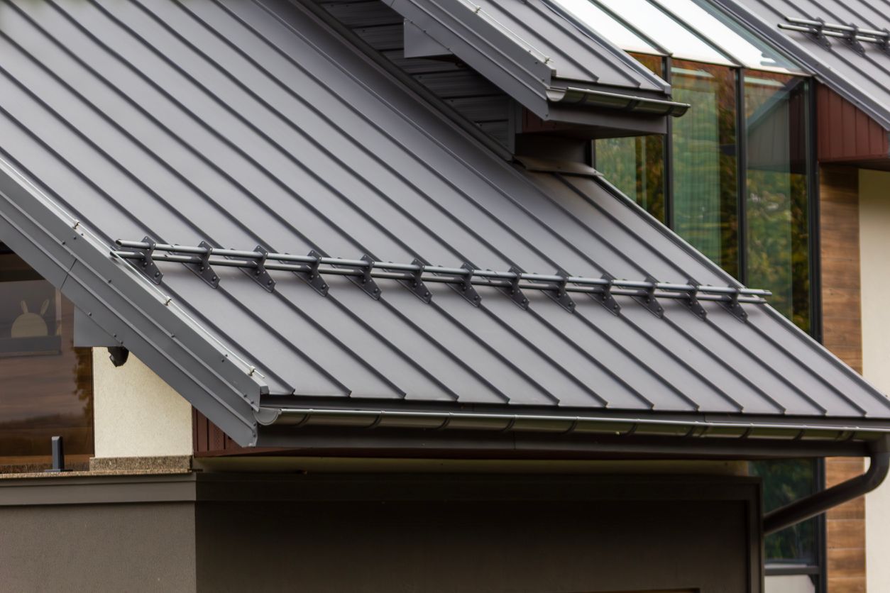 Gray metal roof on a building with a dark gutter and window, cloudy sky.