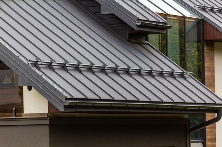 Gray metal roof on a building with a dark gutter and window, cloudy sky.