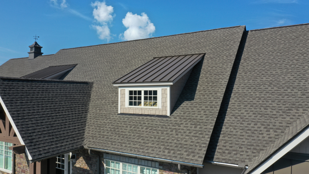 A house roof with shingles and a metal accent roof over a window, against a blue sky.