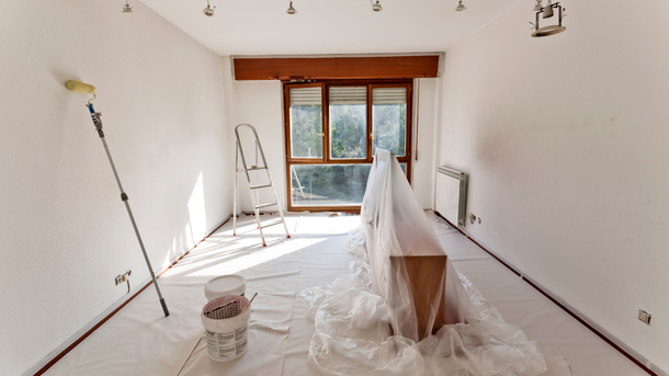 Room being painted white; plastic sheeting covers furniture and floor, paint roller, ladder, sunlight.