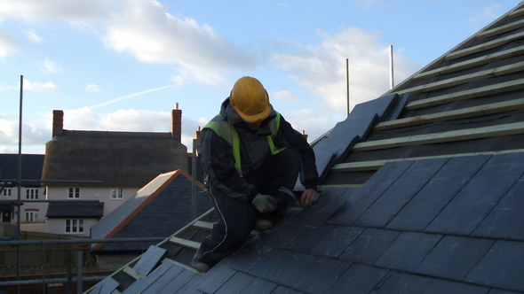 Roofer in a yellow hard hat, kneeling on a roof, installing gray slate tiles. Buildings in the background.