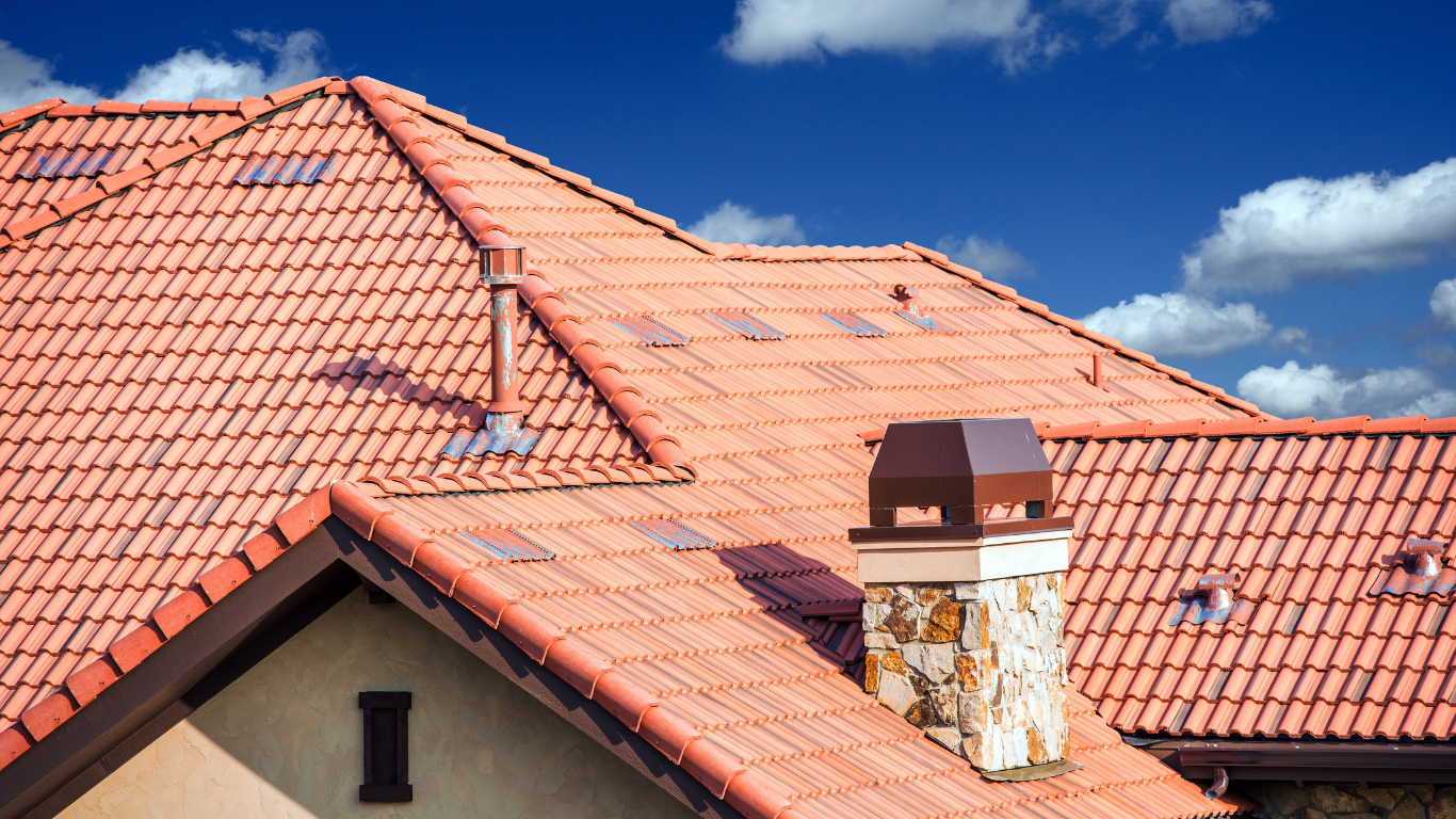 Red tiled roof with chimney and skylights under a blue sky.