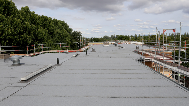 Flat, gray roof with safety rails, vent pipes, and a green tree line under a cloudy sky.