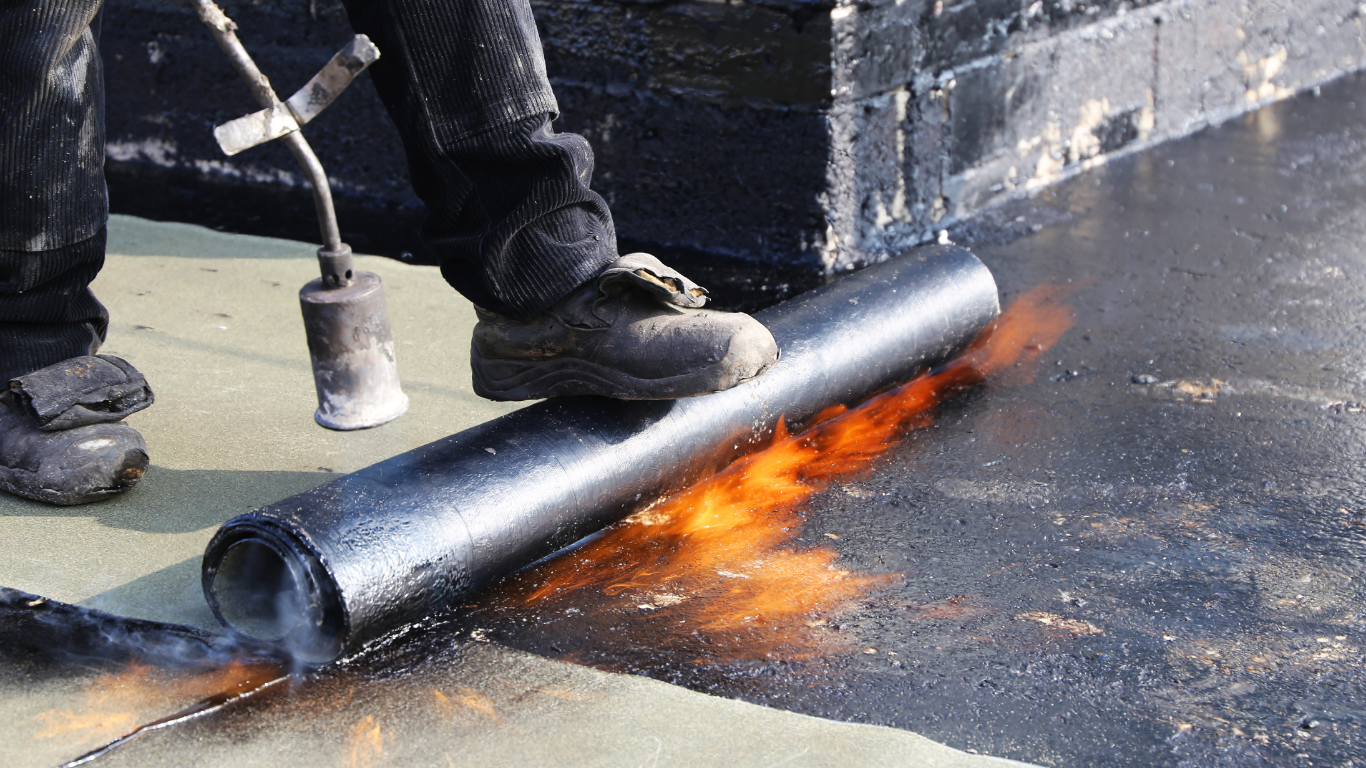Person using a torch to apply roofing material; flames and dark asphalt.