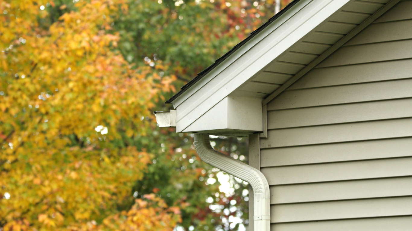 Gutter and downspout on a house with beige siding. Yellow and green autumn leaves in the background.
