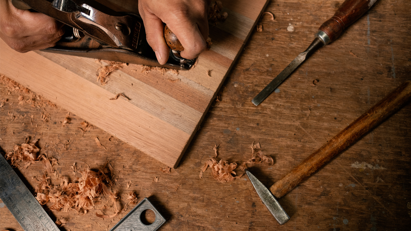 Hands planing wood with a hand plane. Wood shavings, chisel, hammer, and ruler on wooden surface.