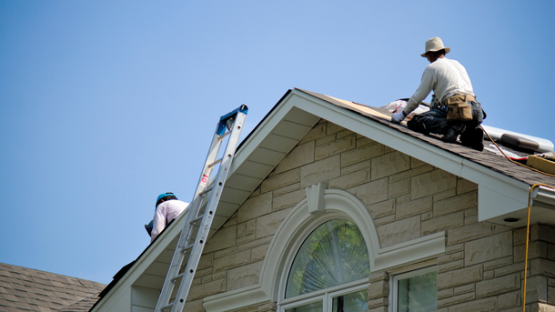 Two roofers working on a shingled roof, sunny day. One on ladder, one installing shingles.