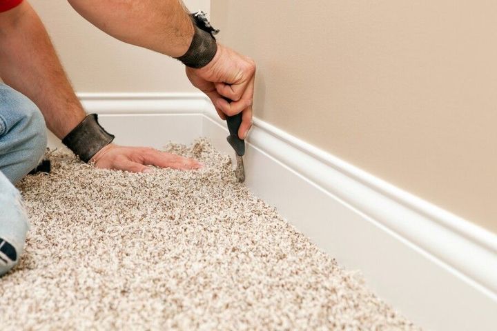 Person using a knife to trim carpet near a white baseboard in a room with tan walls.
