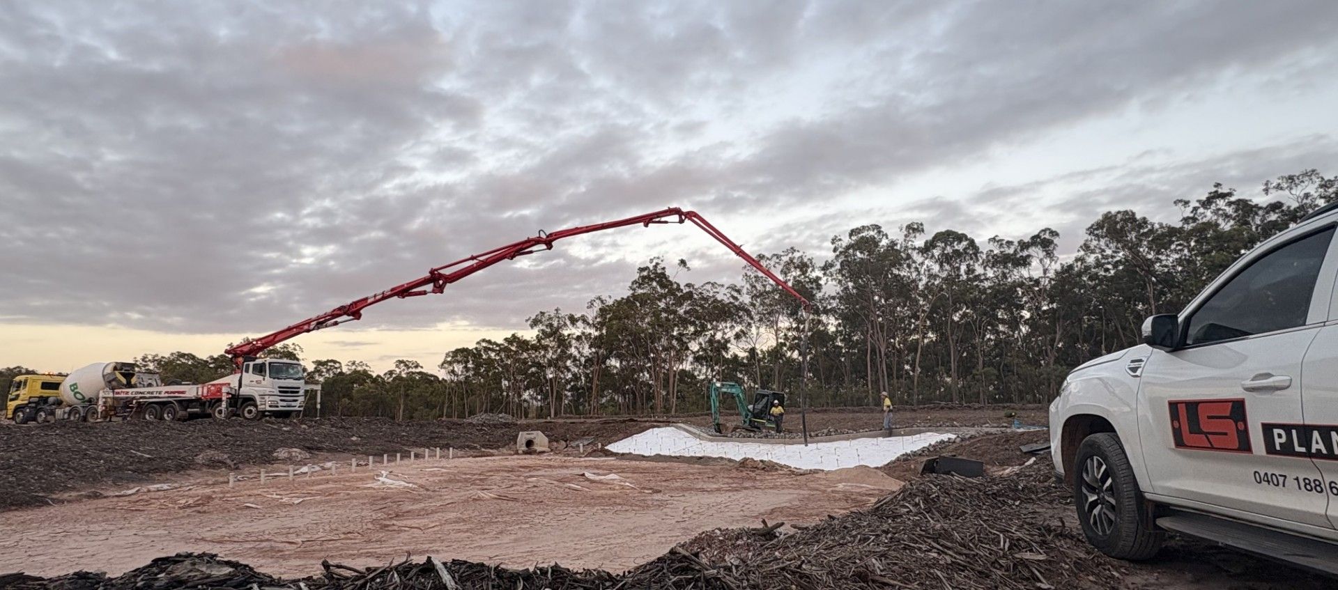 A long-arm concrete pump truck pours concrete onto a construction site at dusk, viewed from behind a white work vehicle. — LS Plant Hire in Calliope, QLD
