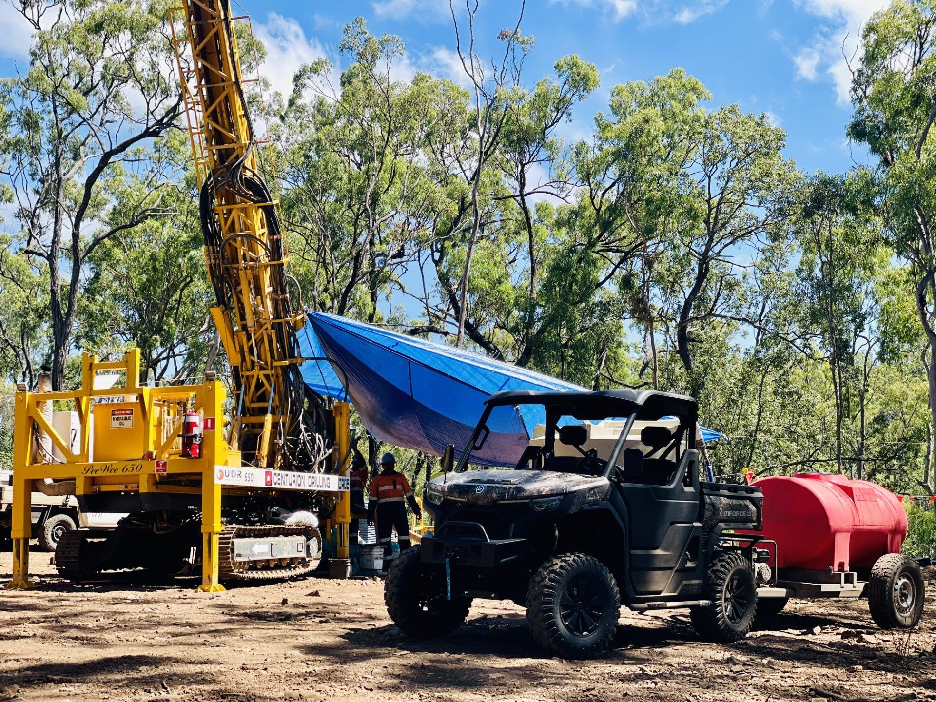 A yellow drill rig operates in a wooded area next to a camouflage ATV pulling a red water tank trailer. — LS Plant Hire in Calliope, QLD
