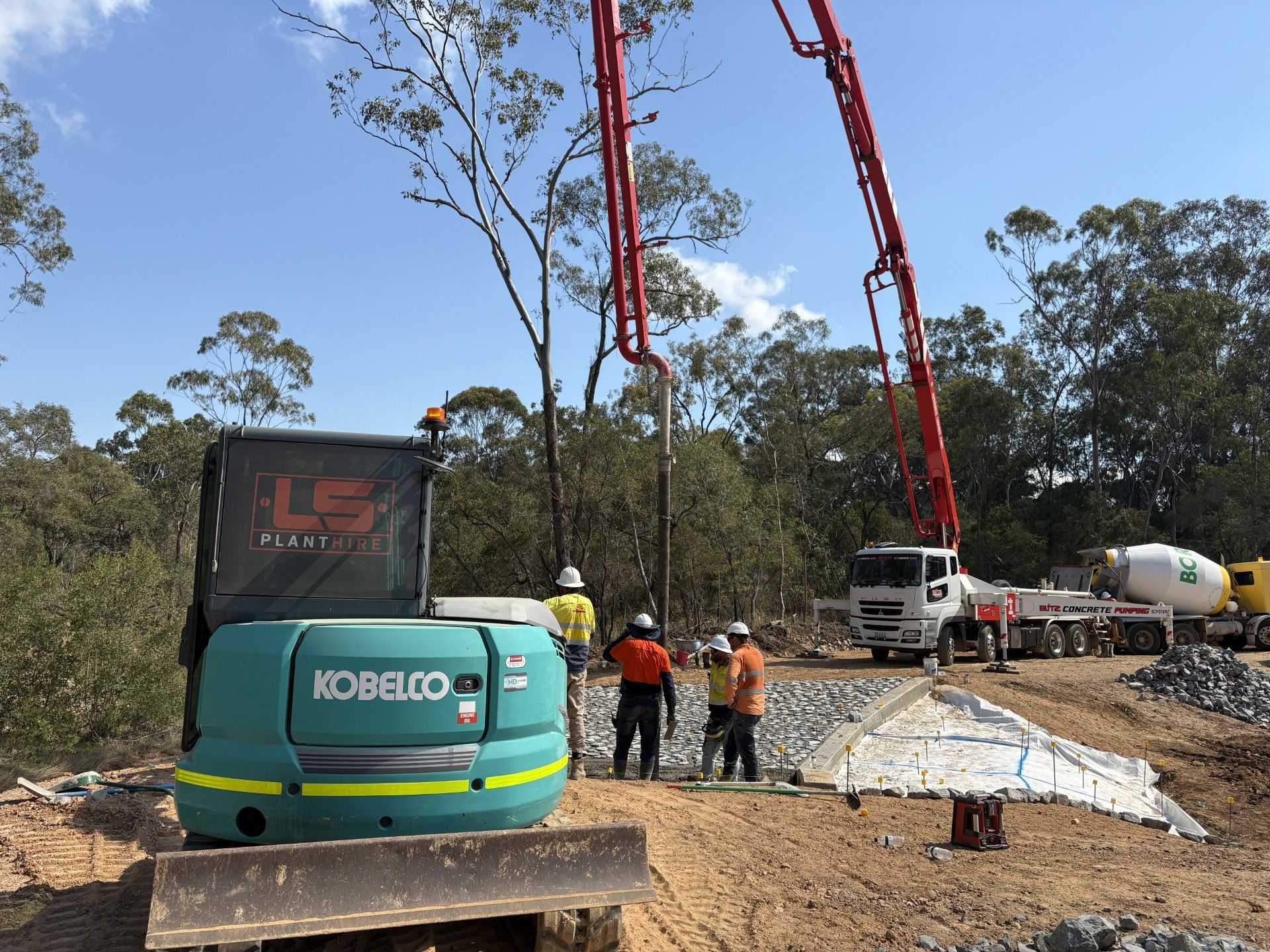 A teal Kobelco excavator sits on a dirt site near construction workers and a concrete pump truck with an extended boom. — LS Plant Hire in Calliope, QLD