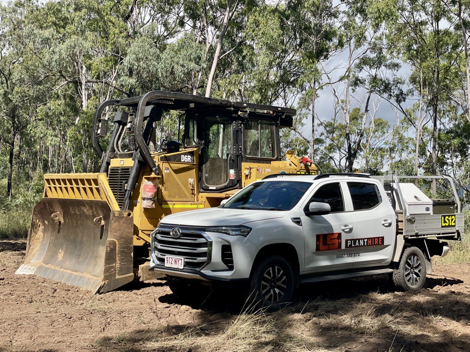 A white pickup truck parked in a dirt clearing next to a large yellow bulldozer in front of a wooded area. — LS Plant Hire in Calliope, QLD