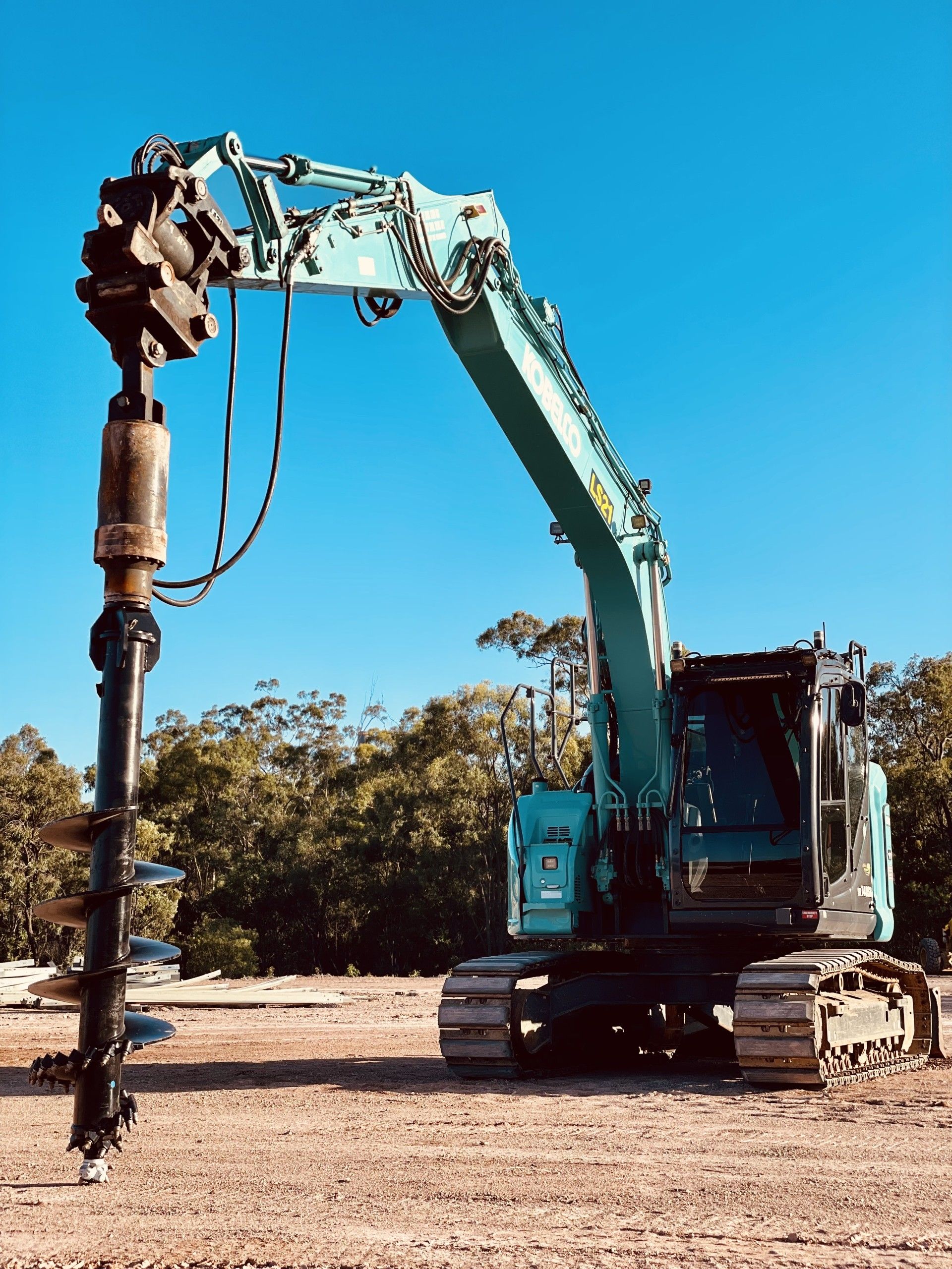 A teal excavator with an attached auger drill bit stands on a dirt construction site against a clear blue sky. — LS Plant Hire in Calliope, QLD