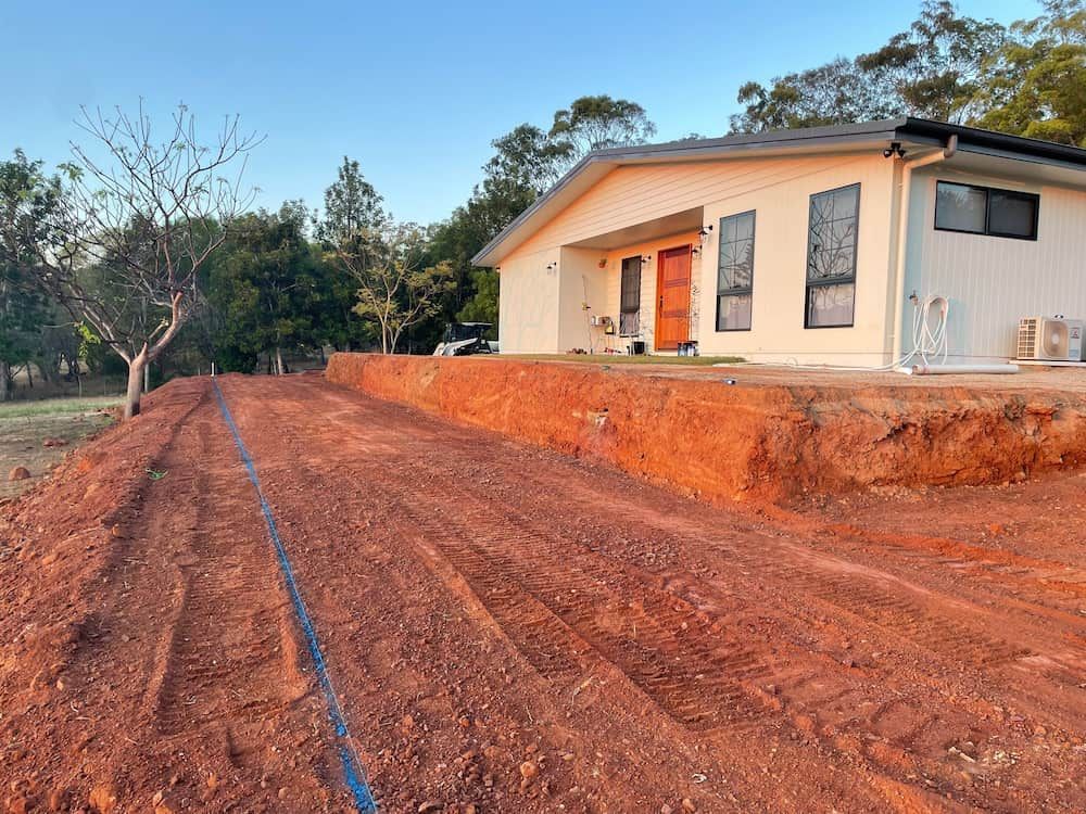 Red Dirt Driveway Leads to a Light-colored House With a Blue Pipe Visible — LS Plant Hire in Calliope, QLD