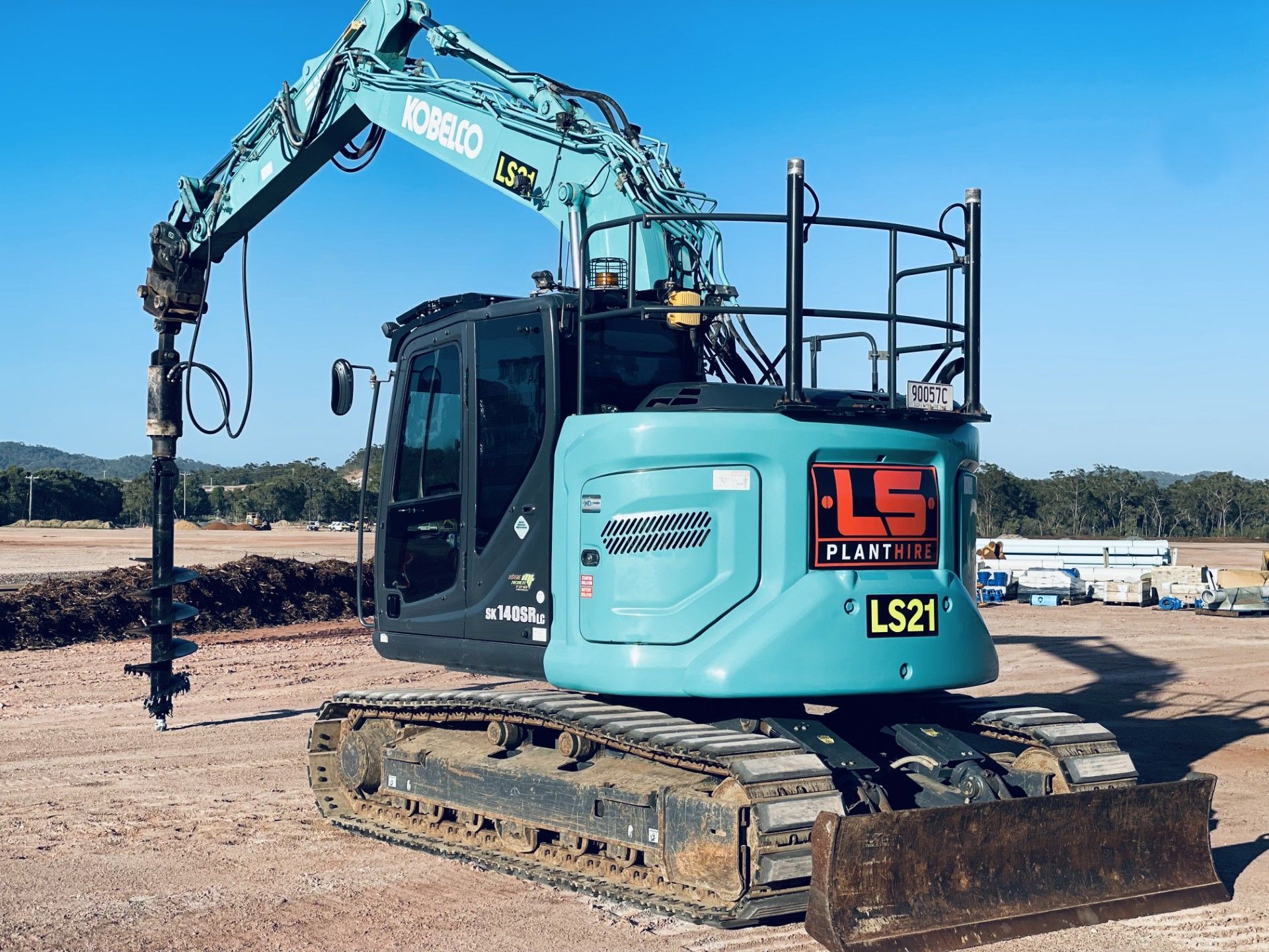 A teal Kobelco excavator with an attached drill auger sits on a dirt construction site under a clear blue sky. — LS Plant Hire in Calliope, QLD