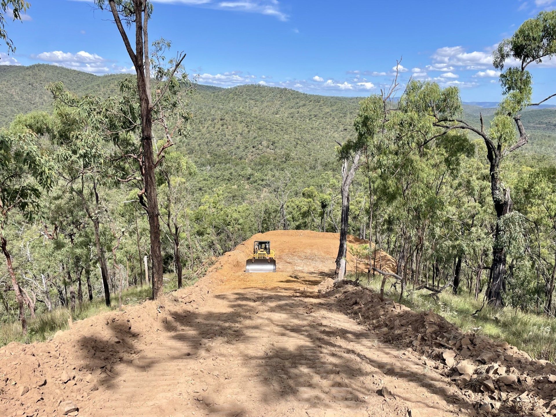 A construction vehicle clears a path through a forested, hilly landscape under a bright blue sky. — LS Plant Hire in Calliope, QLD